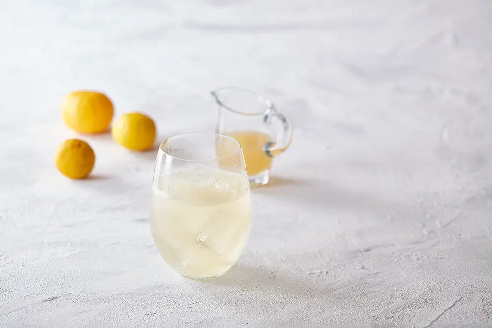 Clear glass with Kayanoya Yuzu Vinegar Drink and a pitcher on a light gray surface with lemons.