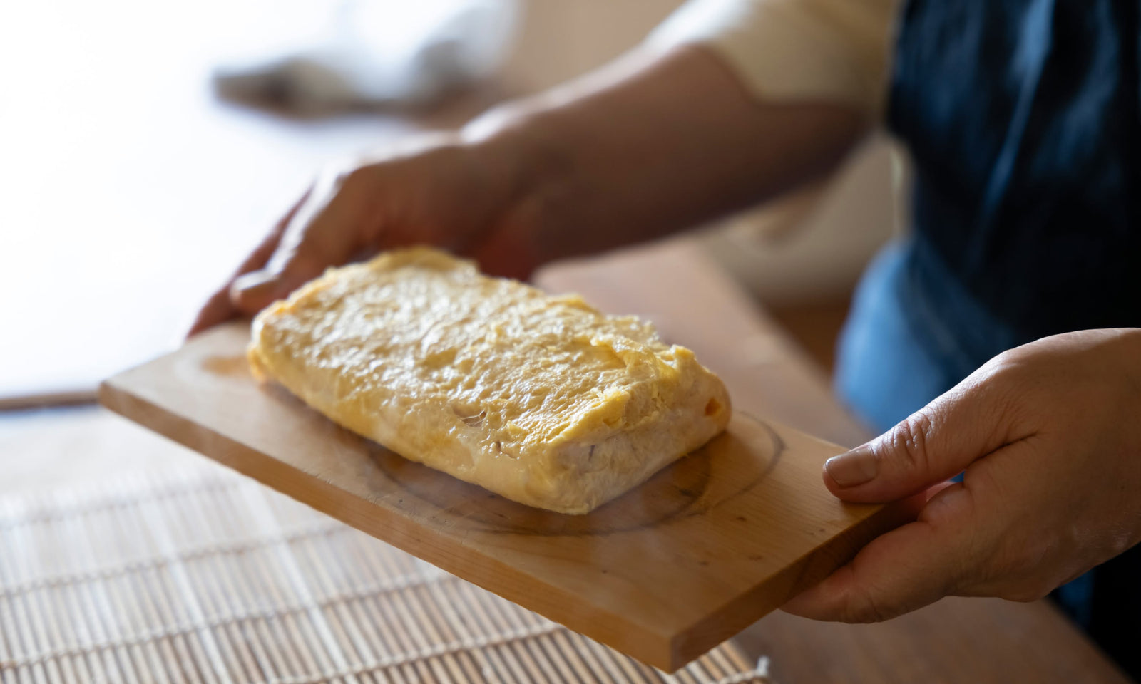 A person holds a wooden board with a freshly cooked Japanese rolled omelet, tamagoyaki, in a kitchen setting.