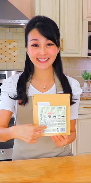 A woman in a light-colored kitchen smiles while holding and pointing to a package with text and images in Japanese. She wears a white shirt and beige apron, standing behind a wooden counter.