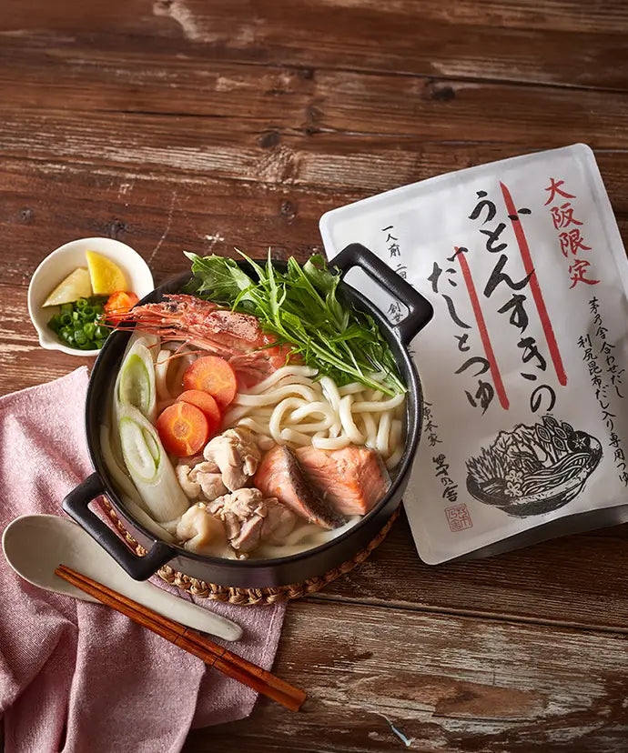 A pot of udon noodle soup with shrimp, chicken, salmon, carrots, scallions, and greens sits on a wooden table beside a packet of soup base, a small bowl of condiments, chopsticks, and a pink napkin.