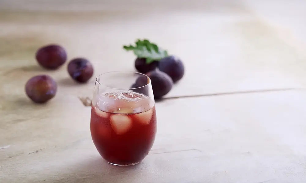 Glass of Kayanoya vinegar drink with ice cubes on a wooden surface with plums around