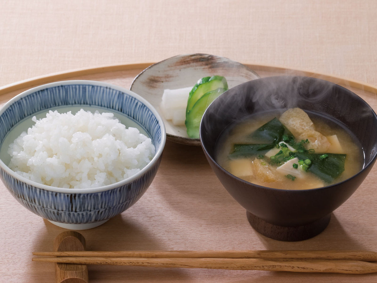 A traditional Japanese meal with a bowl of steamed white rice, a bowl of miso soup with greens and tofu, and a small plate of pickled vegetables, all set on a wooden table with chopsticks.