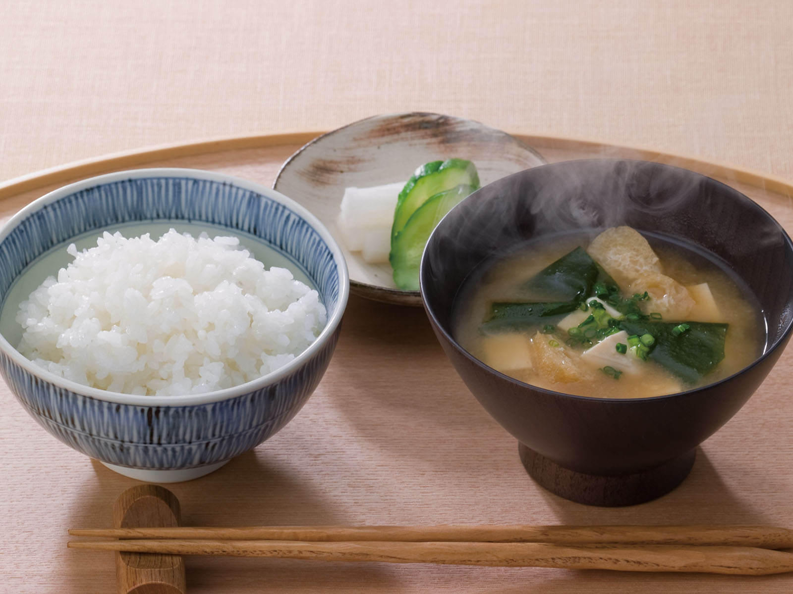 A traditional Japanese meal with a bowl of steamed white rice, a bowl of miso soup with greens and tofu, and a small plate of pickled vegetables, all set on a wooden table with chopsticks.