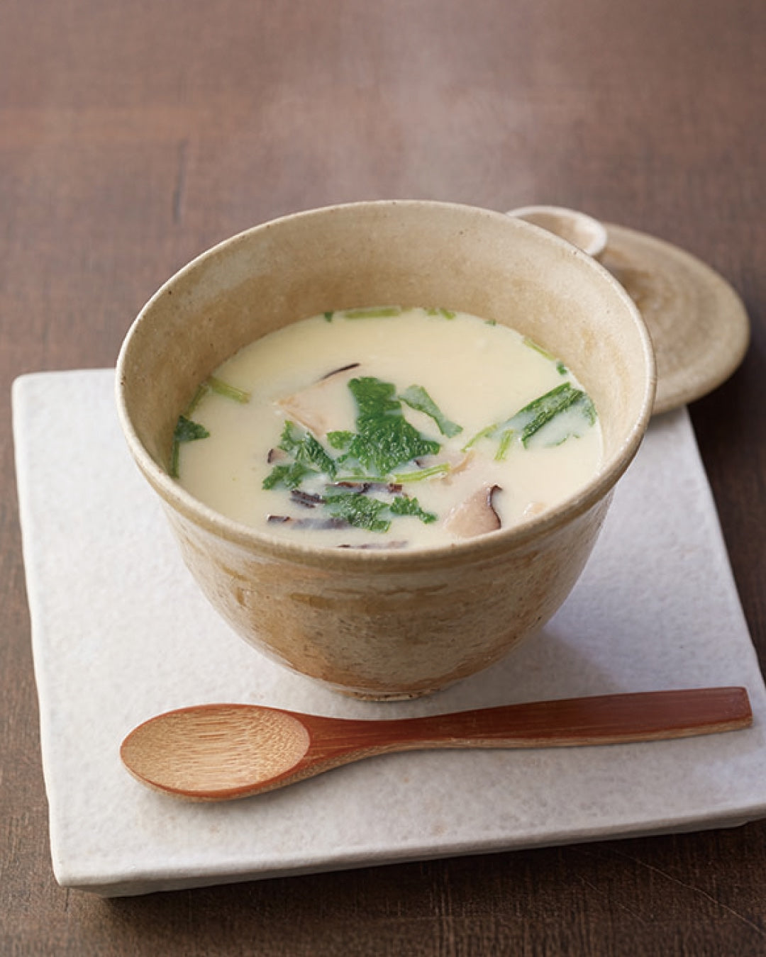 A ceramic bowl filled with creamy soup containing greens and mushroom slices sits on a white square plate with a wooden spoon beside it, all on a wooden table.