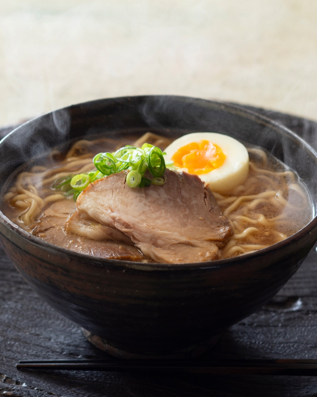 A steaming bowl of ramen topped with sliced pork, a soft-boiled egg, and chopped green onions, placed on a dark wooden surface with black chopsticks beside it.