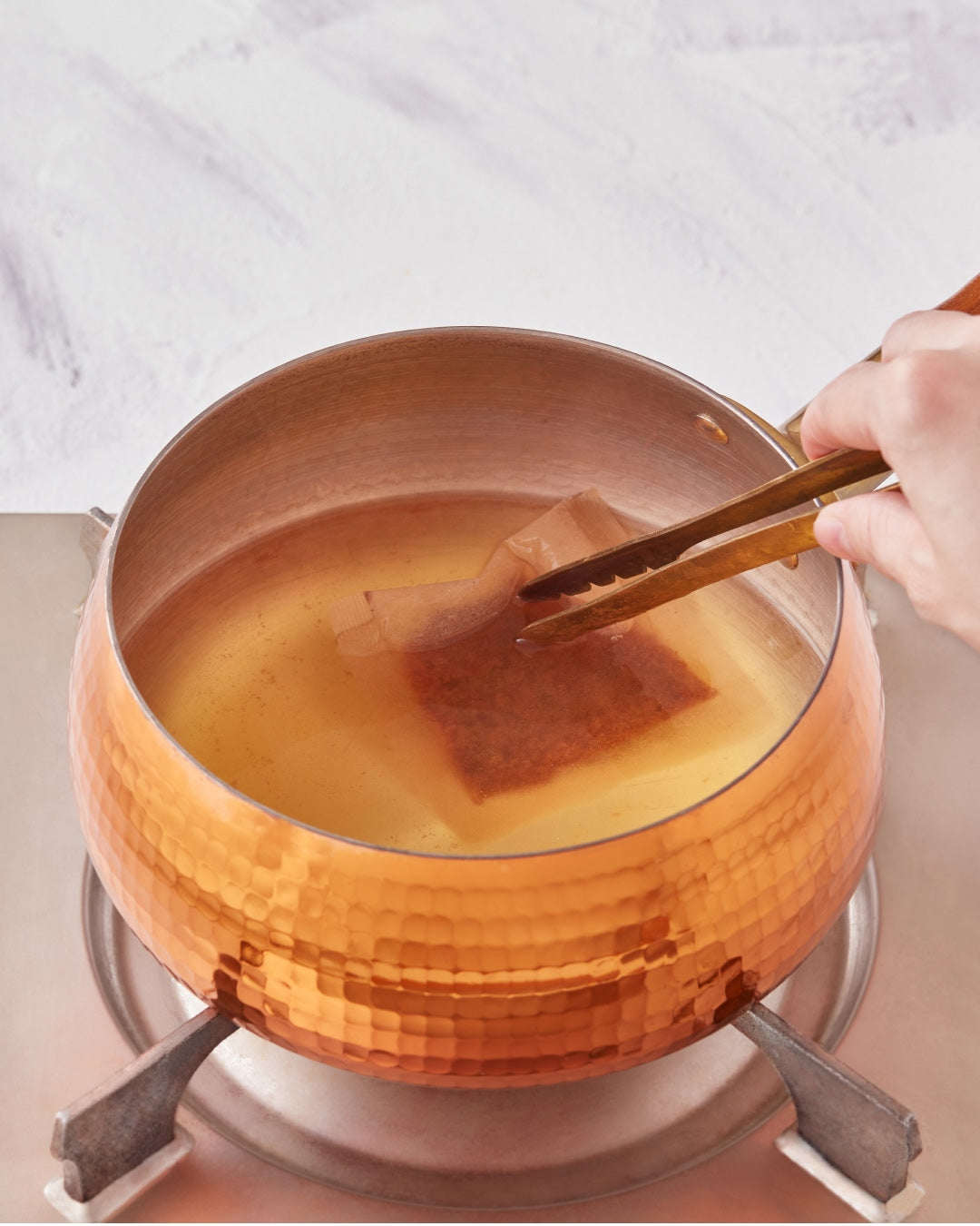 A hand uses tongs to steep a tea bag in a hammered copper pot filled with hot liquid on a stovetop.
