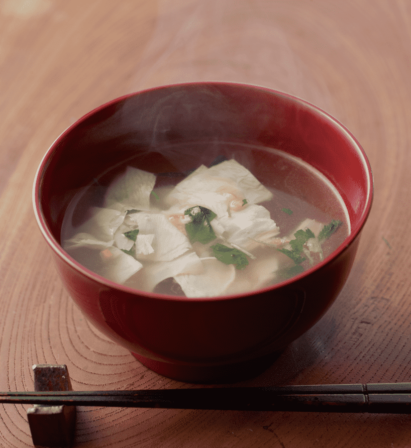 Clear soup with tofu skin and Japanese parsley served in a traditional red lacquer bowl with chopsticks.