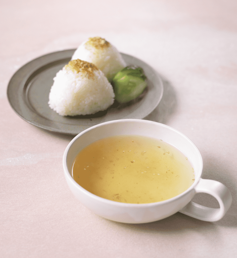A white cup of Dashi Soup Powder - Seafood by Kayanoya USA sits in front, while a plate behind holds two rice balls topped with seasoning, such as skipjack tuna flakes, plus a small side of pickles.