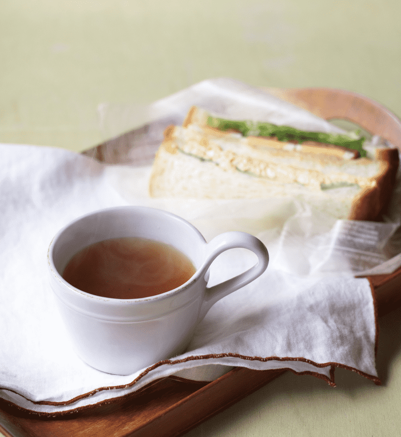 A cup of tea sits on a white napkin beside a sandwich atop a wooden tray with a soft green backdrop, inviting you to enjoy umami-rich flavors with Kayanoya USA's Dashi Soup Powder – Vegetable and Scallop (10 sticks).