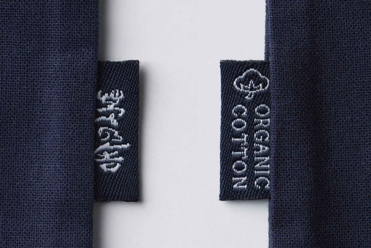 Close-up of two navy blue fabric tags on a Custom Umami Gift Bag - Medium by Kayanoya USA. The left tag has white embroidered script, while the right reads ORGANIC COTTON with a cotton plant icon. Tags are attached to blue garments.