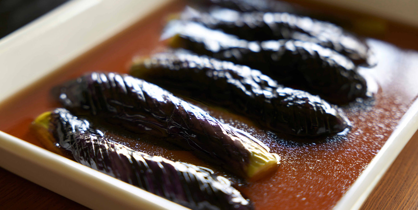 Five cooked Japanese eggplants are arranged in a rectangular white dish, covered in a glossy, brown sauce. The eggplants have a shiny, wrinkled skin and are partially submerged in the sauce.