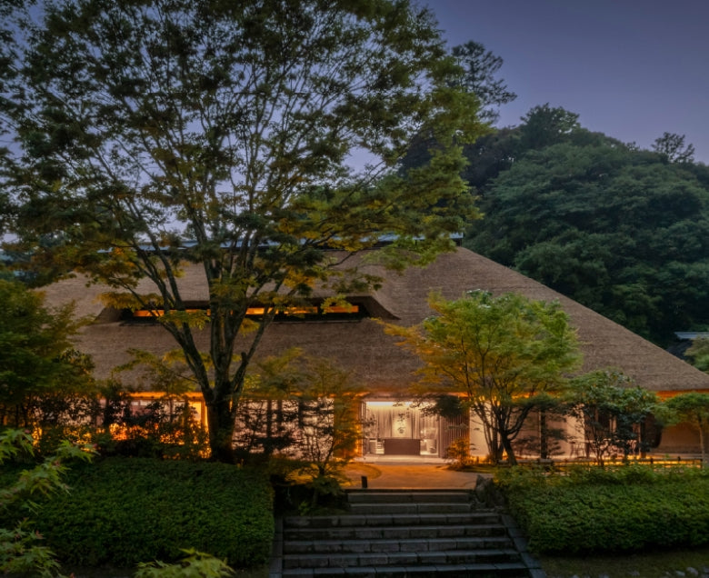 A traditional Japanese-style house with a thatched roof, softly illuminated at dusk, surrounded by lush green trees and plants, with stone steps leading to the entrance.