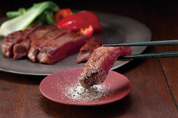 Steak being seasoned with Kayanoya salt on a red plate, with a dark background