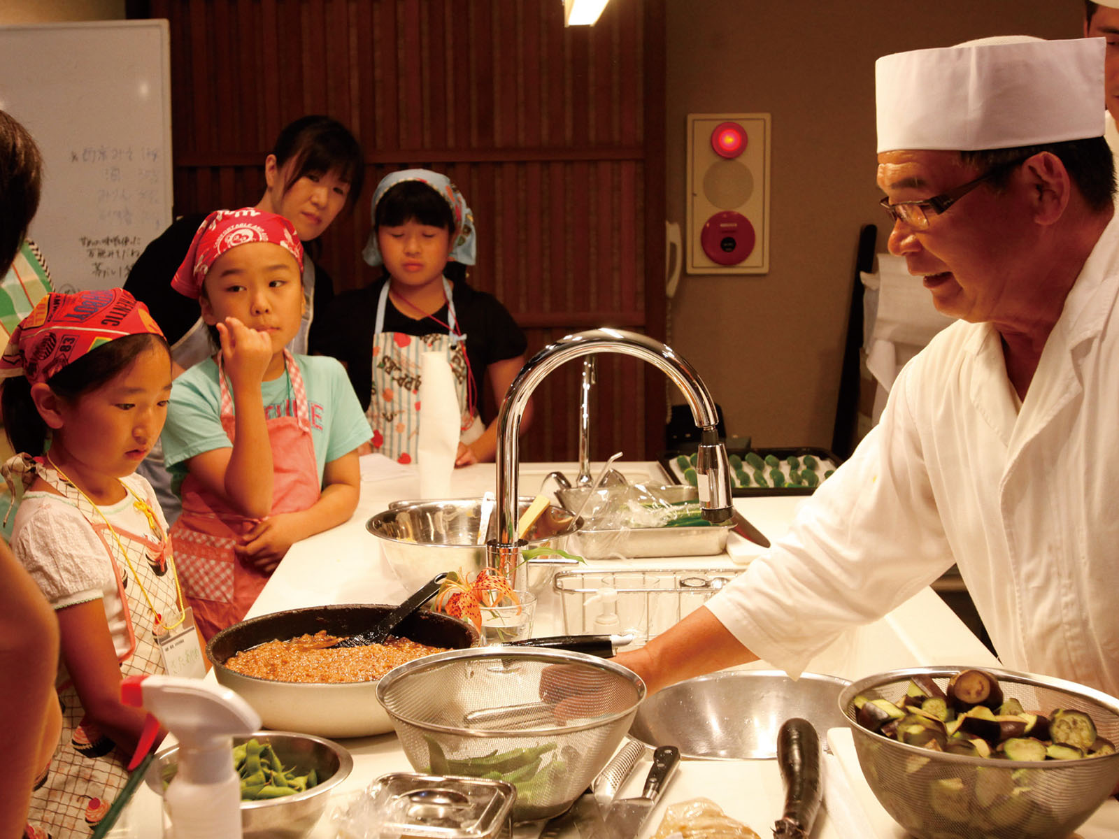 A chef in white attire and hat prepares food at a kitchen counter, while a group of children and adults watch attentively. Various ingredients and cooking utensils are visible on the counter.