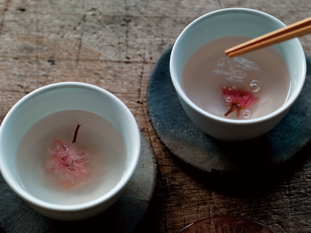 Two white cups containing a clear liquid with a pink cherry blossom flower inside each, placed on round coasters on a wooden surface. A pair of chopsticks hovers above one of the cups.