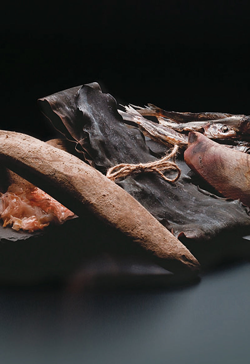 A still life composition of traditional Japanese ingredients, including a piece of dried kombu seaweed, a dried fish tied with string, shaved bonito flakes, and a stick of dried bonito, set against a dark background.