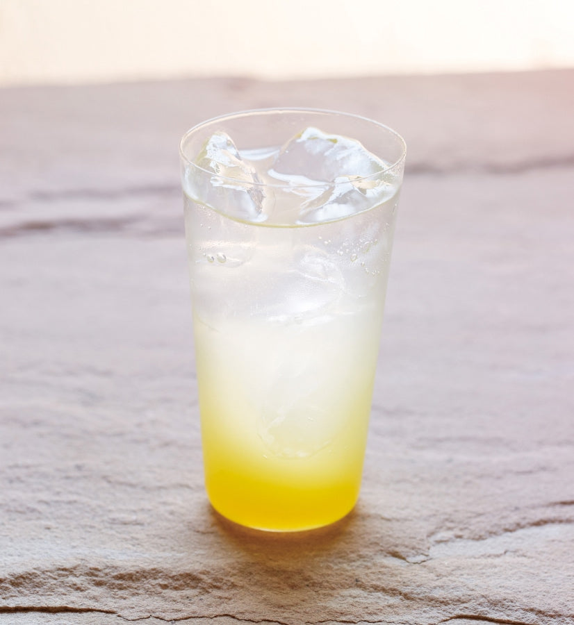 A clear glass with ice holds a yellow gradient drink made from [Seasonal] Vinegar Drink Concentrate - Shikuwasa (150 ml) by Kayanoya USA - Kubara Honke USA, Inc., its color lighter at the top and deeper at the bottom on a light textured surface.
