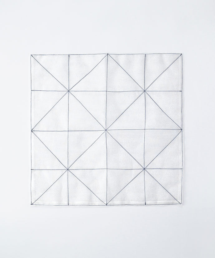 The Kayanoya Custom-Made 'Aya' Dish Towel by Kayanoya USA features a white square cloth with intersecting black lines forming geometric triangles, created in collaboration with Shirayuki-Fukin. The background is plain and light-colored.
