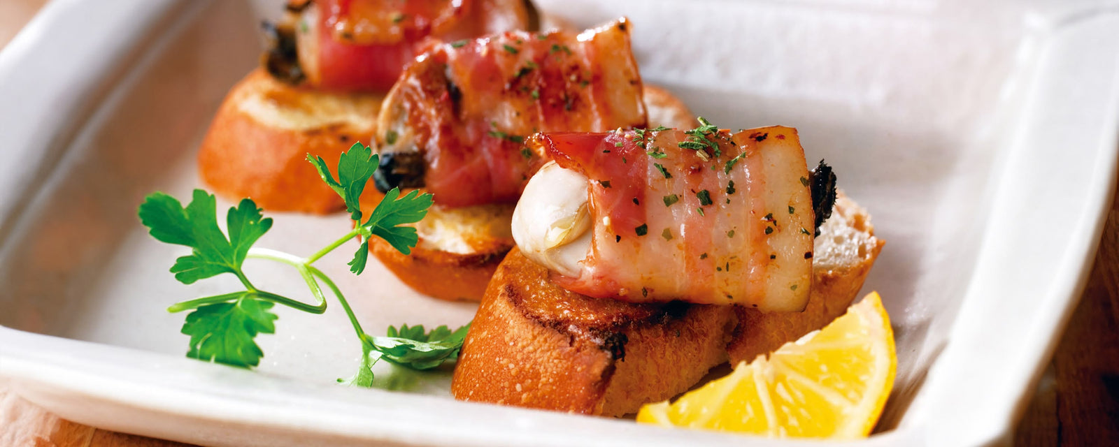 Grilled bacon-wrapped oysters on toasted bread with herbs and a lemon wedge.