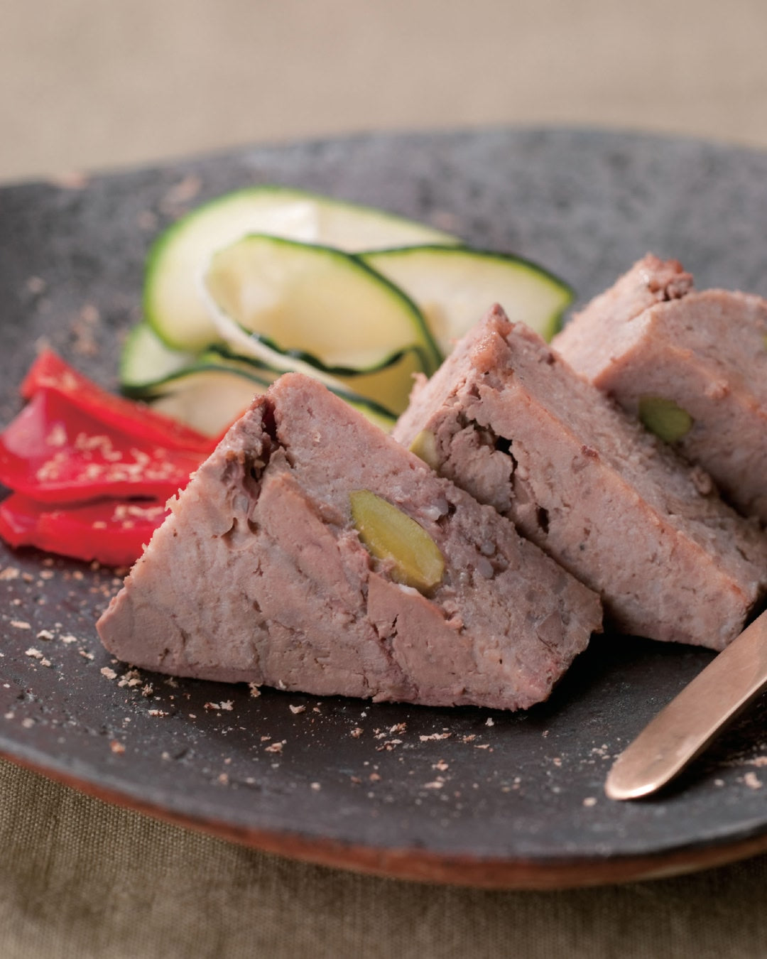 Slices of savory meat pate studded with pistachios, served on a dark plate with red pepper strips and thinly sliced zucchini in the background, lightly sprinkled with seasoning.