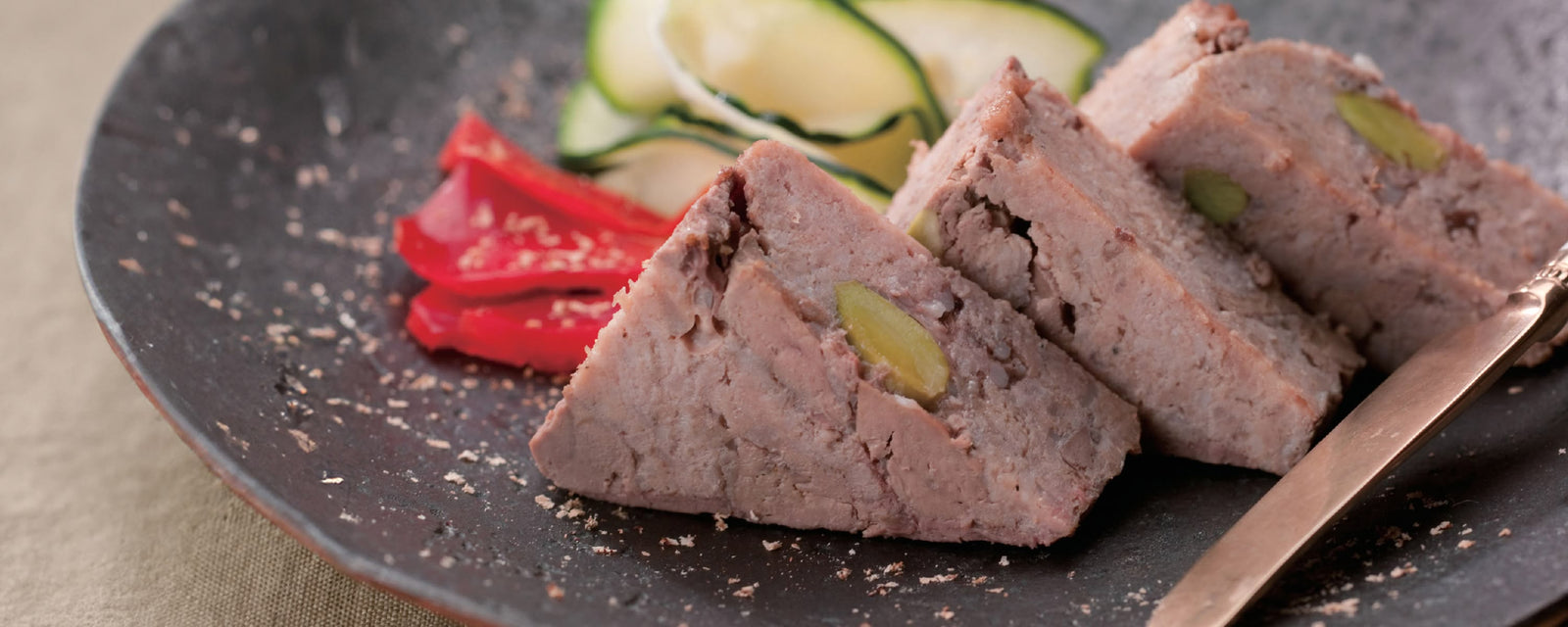 Slices of savory meat pate studded with pistachios, served on a dark plate with red pepper strips and thinly sliced zucchini in the background, lightly sprinkled with seasoning.