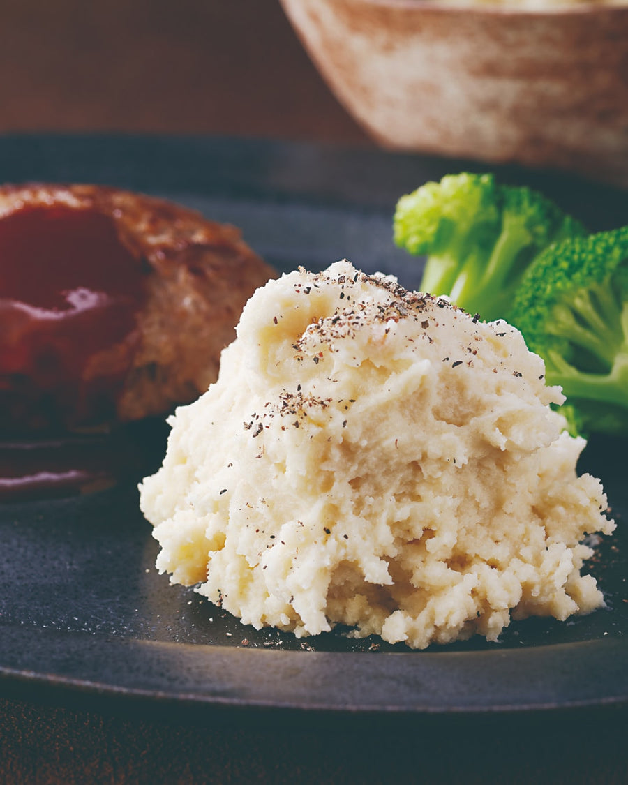 A serving of creamy mashed potatoes topped with cracked black pepper, plated alongside a hamburger steak with glossy brown sauce and steamed broccoli on a dark plate.