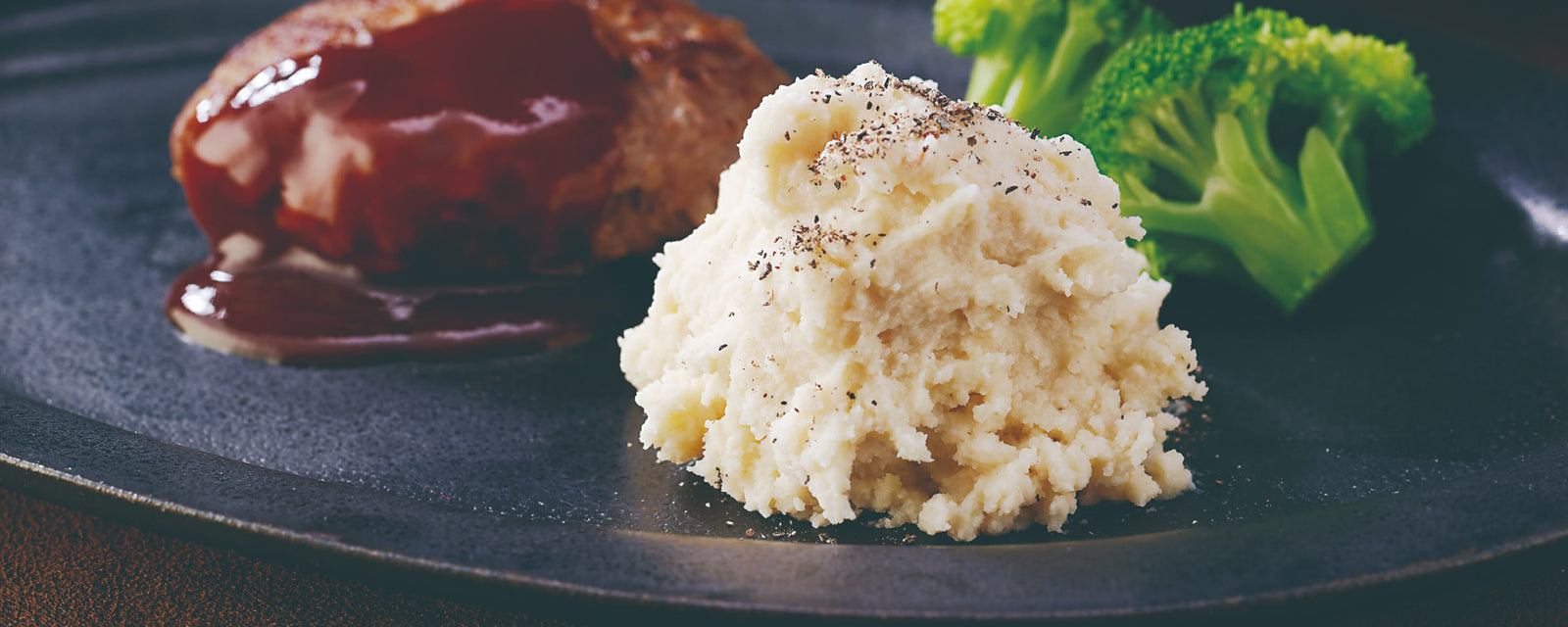 A serving of creamy mashed potatoes topped with cracked black pepper, plated alongside a hamburger steak with glossy brown sauce and steamed broccoli on a dark plate.