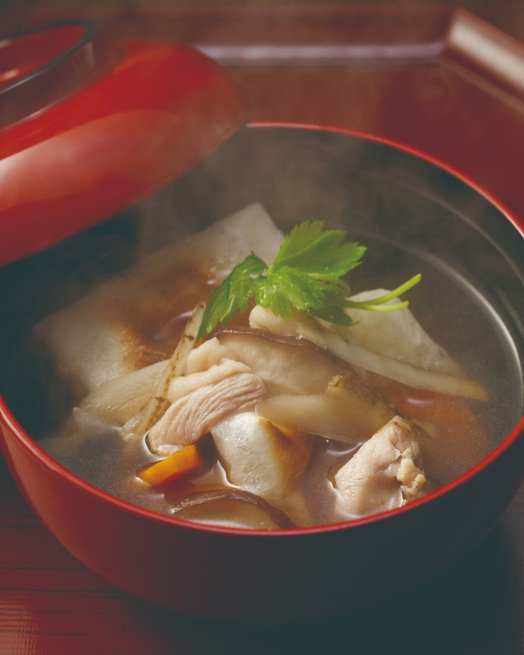 A red lacquer bowl of steaming Japanese ozoni soup with toasted mochi, chicken slices, mushrooms, and vegetables in a clear dashi broth, garnished with fresh mitsuba leaves.