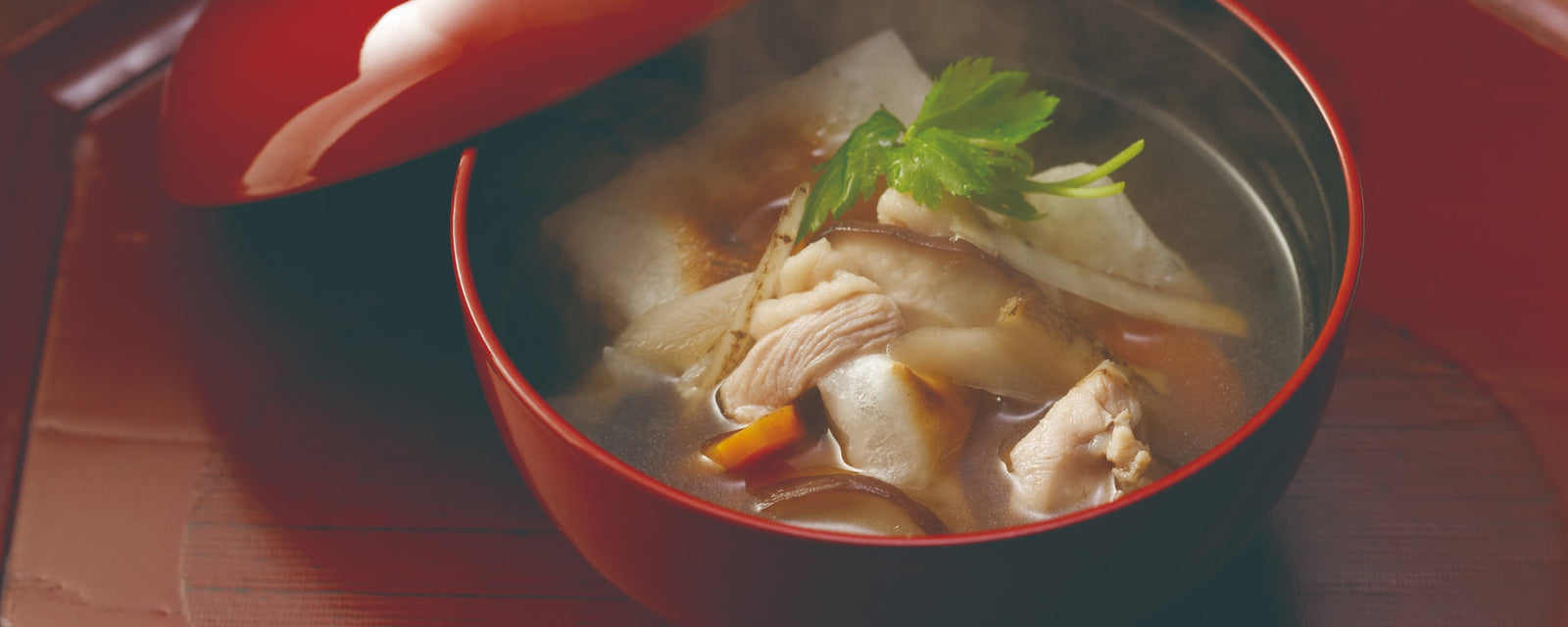A red lacquer bowl of steaming Japanese ozoni soup with toasted mochi, chicken slices, mushrooms, and vegetables in a clear dashi broth, garnished with fresh mitsuba leaves.