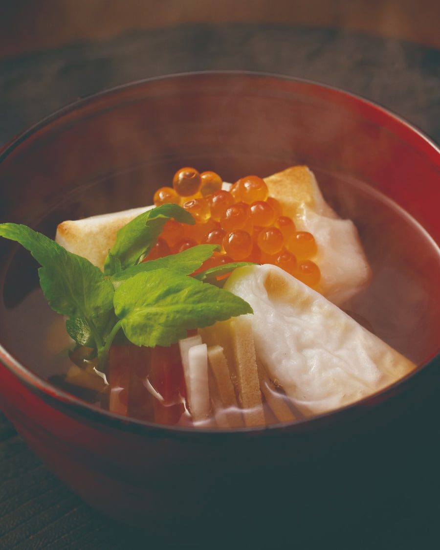 A red lacquer bowl of steaming Japanese ozoni soup with toasted mochi rice cakes, salmon roe (ikura), and seri (Japanese parsley) leaves in a clear dashi broth.