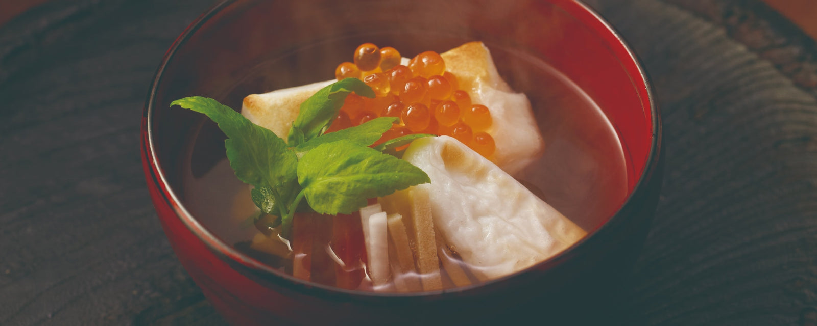 A red lacquer bowl of steaming Japanese ozoni soup with toasted mochi rice cakes, salmon roe (ikura), and seri (Japanese parsley) leaves in a clear dashi broth.