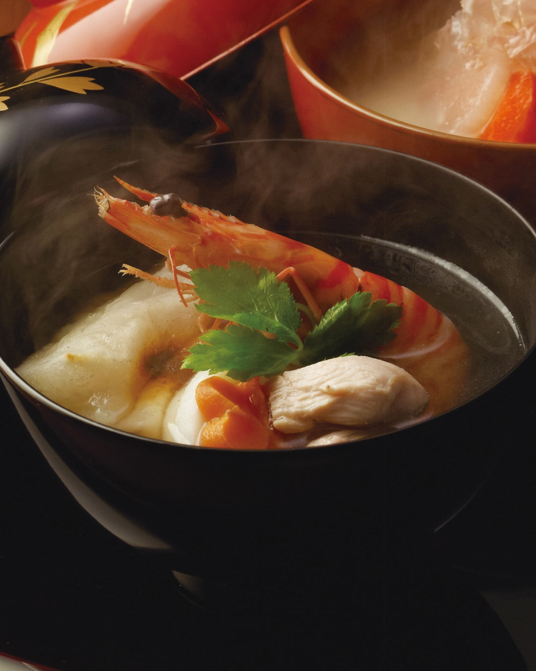 A black lacquer bowl of steaming Japanese ozoni soup with a toasted mochi rice cake, shrimp, chicken, carrot, and fresh mitsuba leaves in a clear broth.