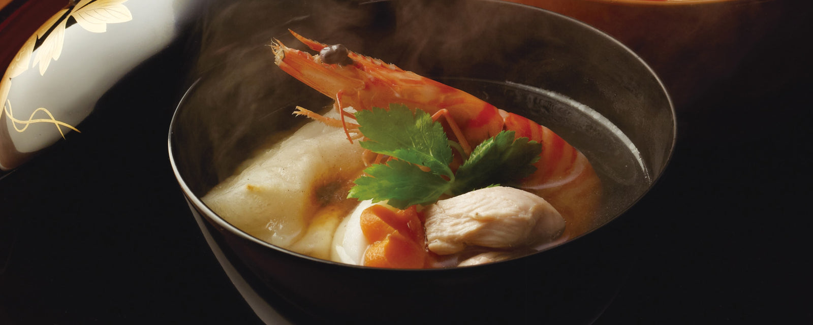 A black lacquer bowl of steaming Japanese ozoni soup with a toasted mochi rice cake, shrimp, chicken, carrot, and fresh mitsuba leaves in a clear broth.