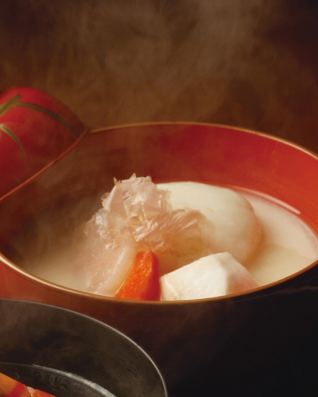 A red lacquer bowl of steaming Japanese soup with a large round mochi rice cake, taro potato, daikon radish, and a slice of carrot in a light broth, with the decorative lid partially open.