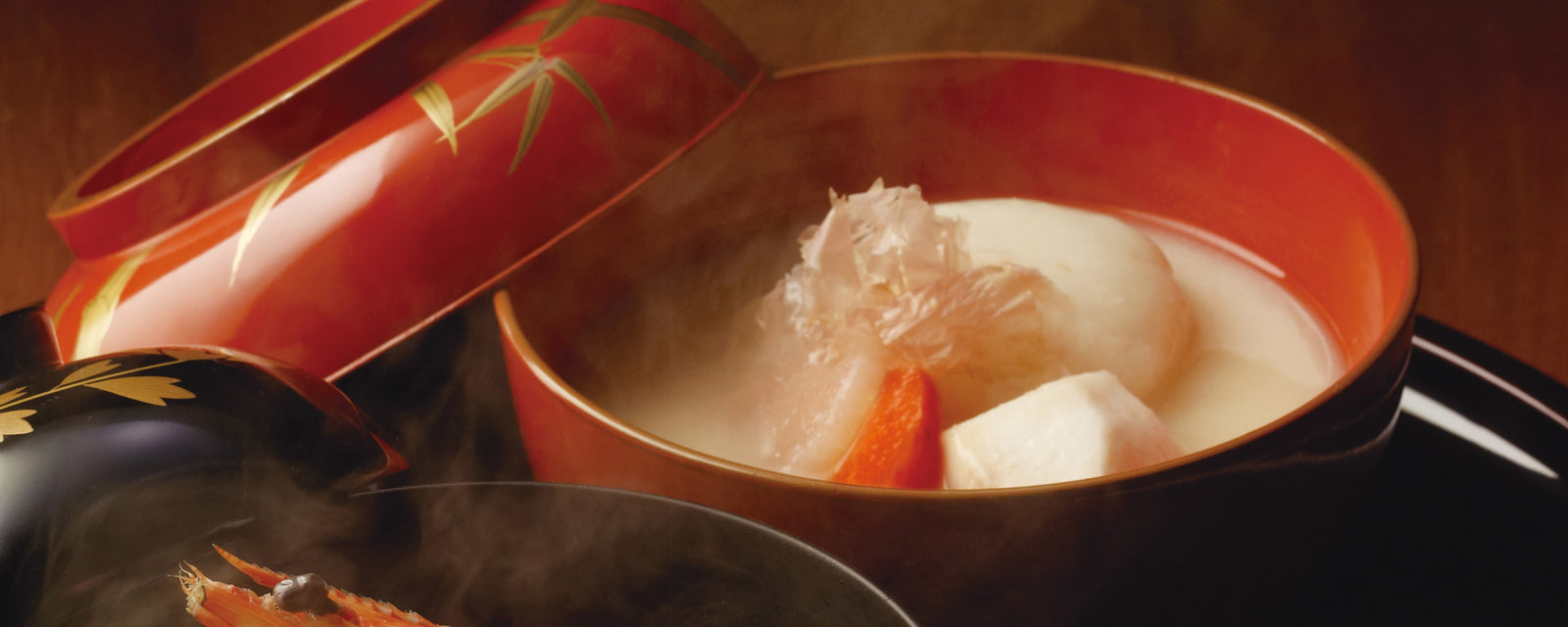A red lacquer bowl of steaming Japanese soup with a large round mochi rice cake, taro potato, daikon radish, and a slice of carrot in a light broth, with the decorative lid partially open.