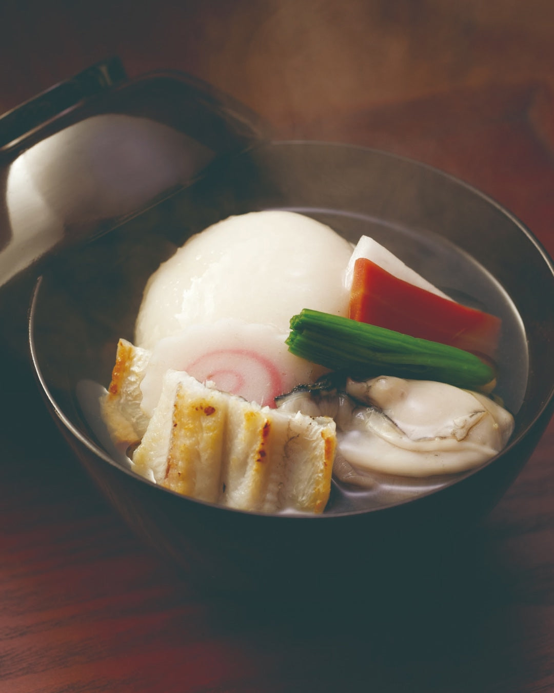 A black bowl of clear Japanese soup with a large round mochi rice cake, grilled fish cake slices, a piece of sea eel, an oyster, and colorful vegetables like carrot and greens, served in a light broth.