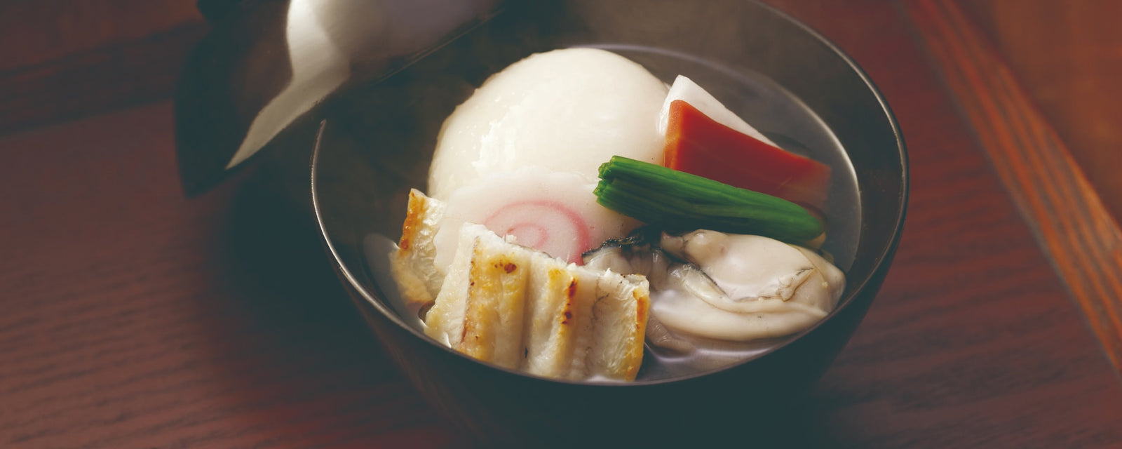 A black bowl of clear Japanese soup with a large round mochi rice cake, grilled fish cake slices, a piece of sea eel, an oyster, and colorful vegetables like carrot and greens, served in a light broth.