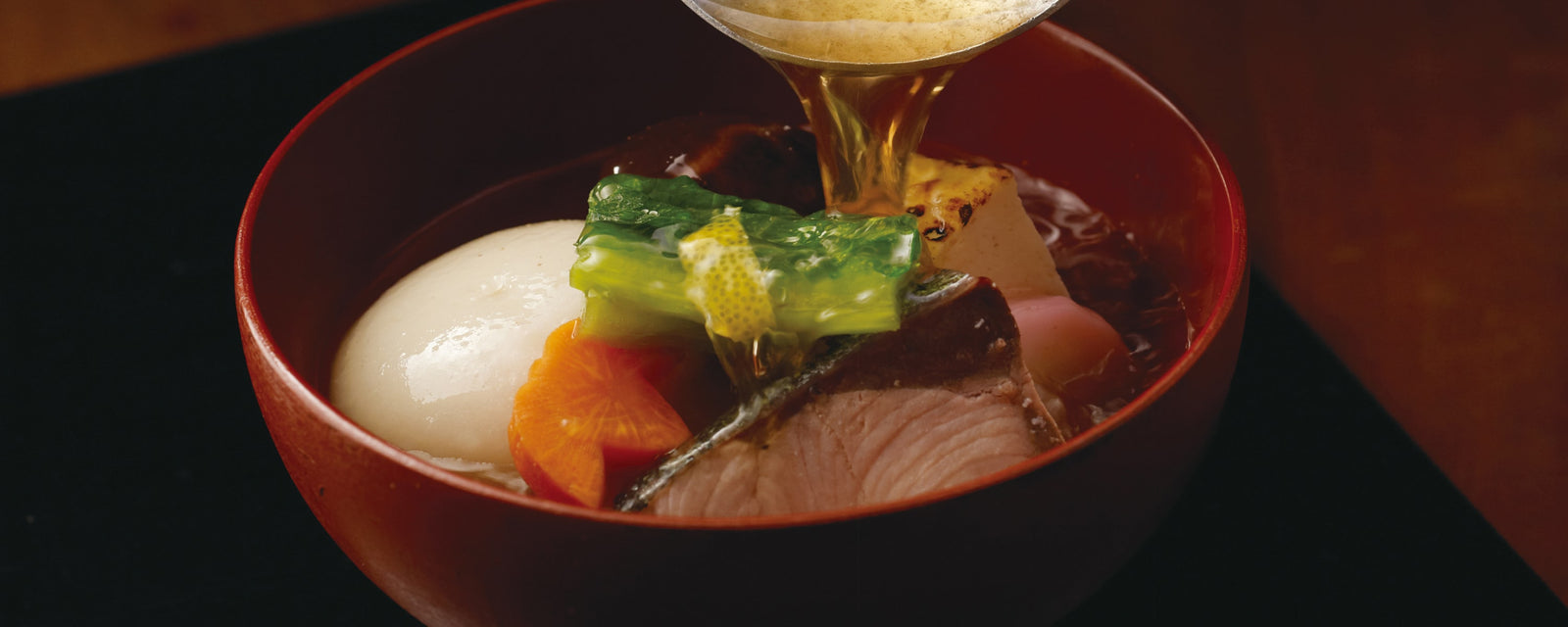 A lacquered red bowl of traditional Japanese soup with assorted ingredients—round mochi, sliced carrot, grilled tofu, greens, and a piece of fish—while clear hot broth is being poured over the top.