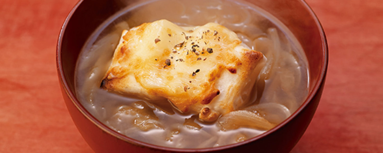 A bowl of Japanese onion soup topped with a toasted mochi square, lightly browned and sprinkled with black pepper, served in a lacquered bowl.