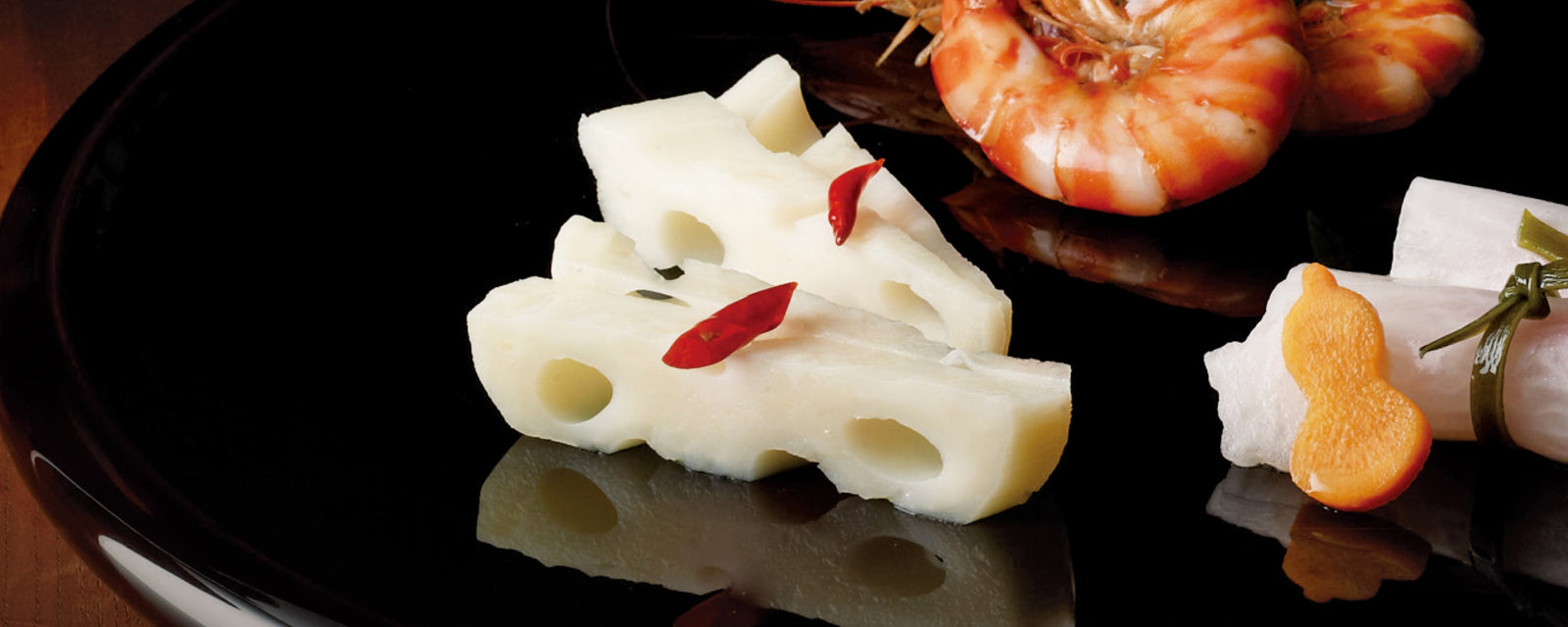 Slices of lotus root topped with small red chili pieces, served on a glossy black plate alongside shrimp.