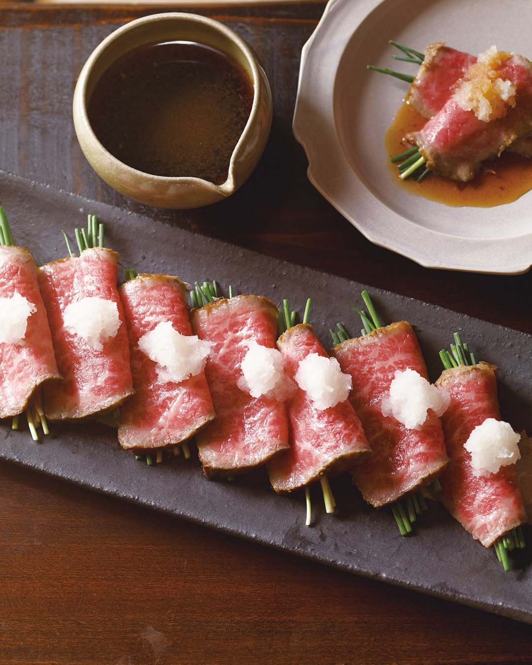Thin slices of seared beef rolled around green onions, topped with small mounds of grated daikon radish, arranged on a rectangular black plate.