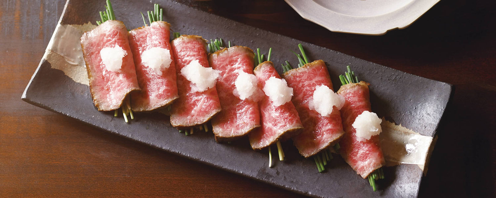 Thin slices of seared beef rolled around green onions, topped with small mounds of grated daikon radish, arranged on a rectangular black plate.