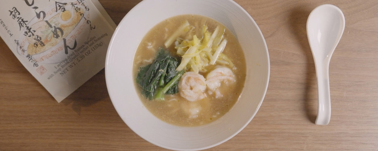 Ramen with creamy light-colored soup in a white bowl, topped with shrimp, leafy greens, and sliced leeks, served on a wooden table with a white spoon.