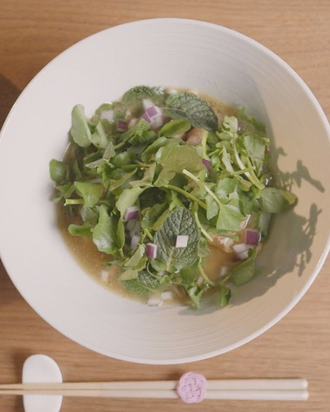 A light herbal soup in a white bowl, topped with fresh greens, mint leaves, and diced red onion, served on a wooden table with a white spoon.
