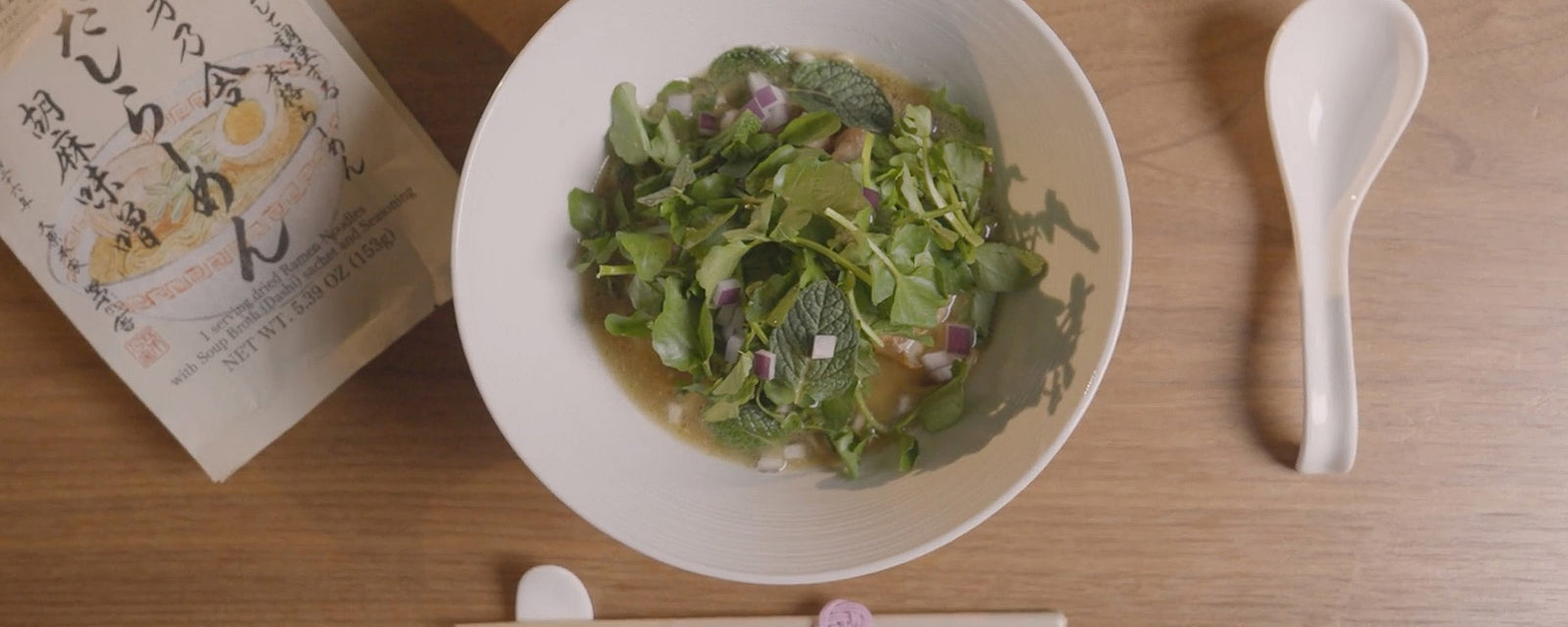 A light herbal soup in a white bowl, topped with fresh greens, mint leaves, and diced red onion, served on a wooden table with a white spoon.
