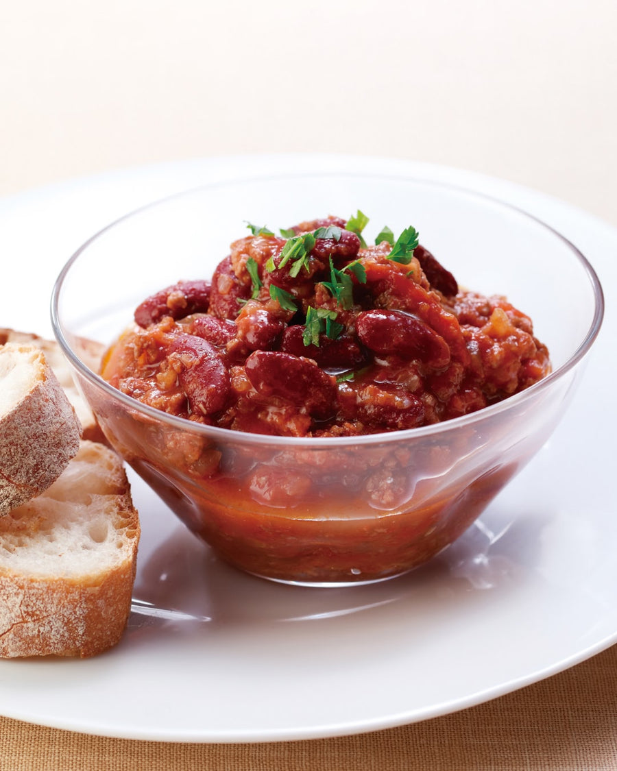 A glass bowl of hearty kidney bean stew in a rich tomato sauce, garnished with fresh herbs, served with slices of crusty bread on a white plate.