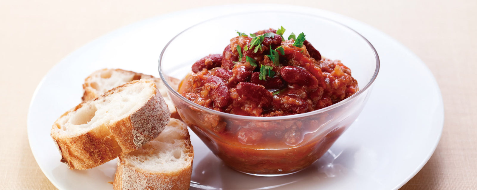 A glass bowl of hearty kidney bean stew in a rich tomato sauce, garnished with fresh herbs, served with slices of crusty bread on a white plate.