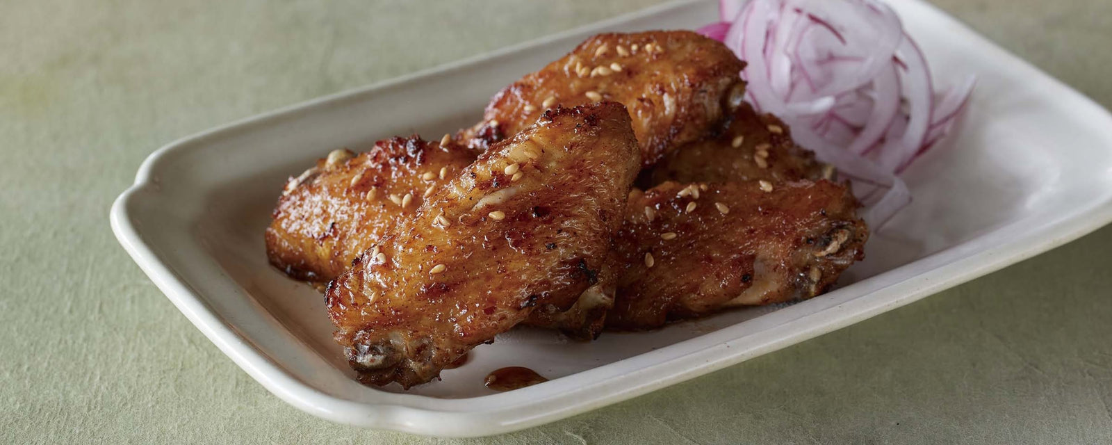 Glazed chicken wings sprinkled with sesame seeds, served on a rectangular plate with thinly sliced red onions on the side.