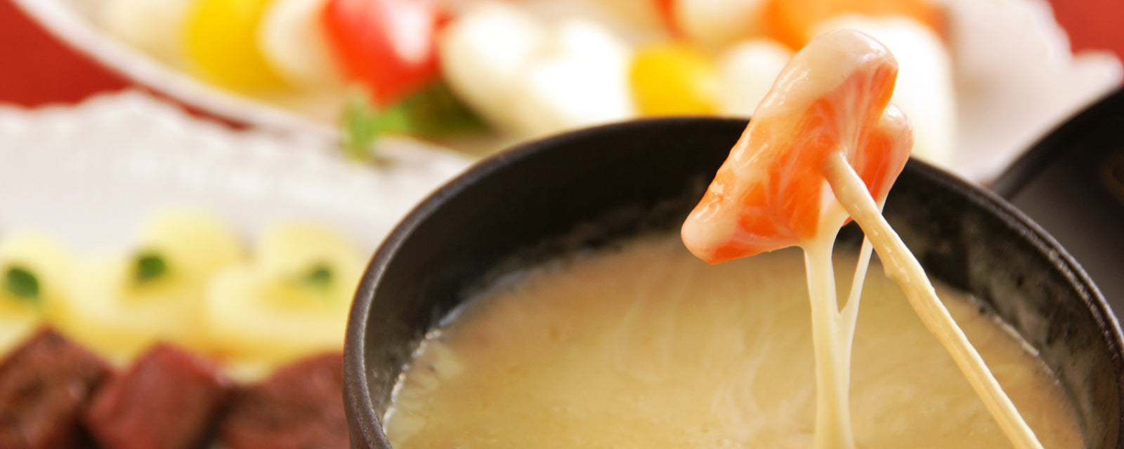 A piece of carrot being dipped into a pot of creamy melted cheese fondue, with colorful vegetables in the background.