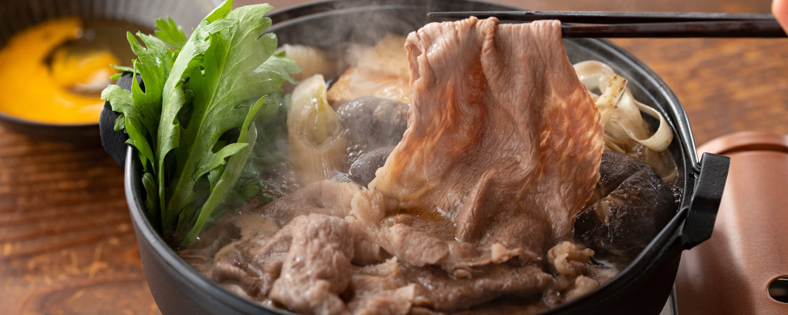 Thinly sliced beef being lifted with chopsticks from a steaming hot pot, surrounded by leafy greens, mushrooms, and leeks in a bubbling broth.